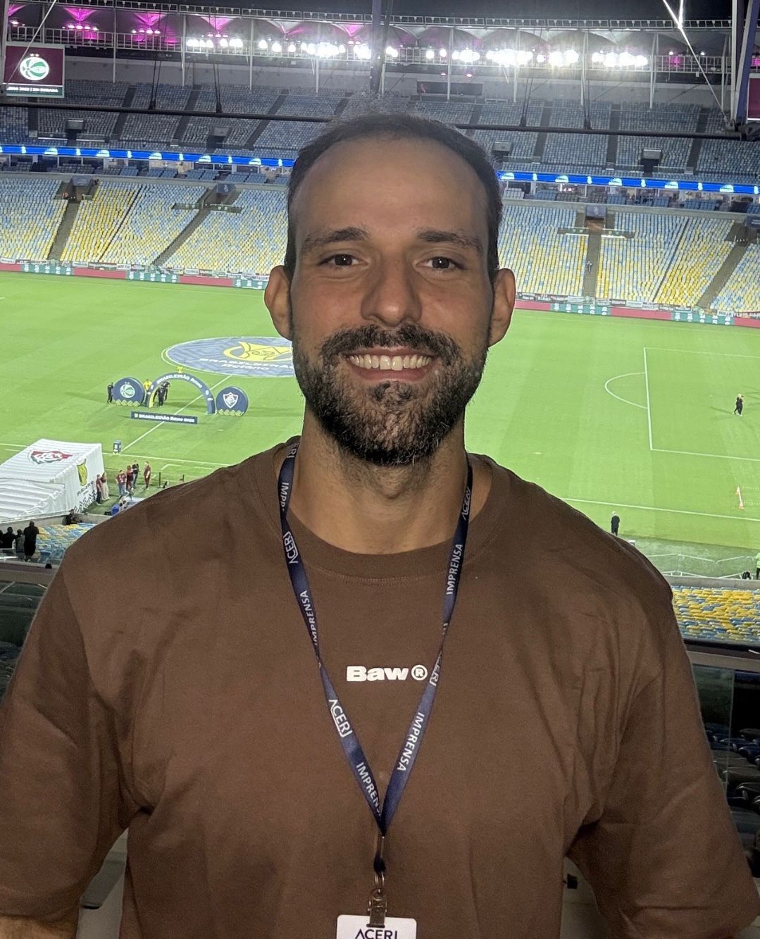 Bernardo Couto no estádio à noite, sorrindo, com credencial de imprensa ACERJ; arquibancadas e gramado ao fundo
